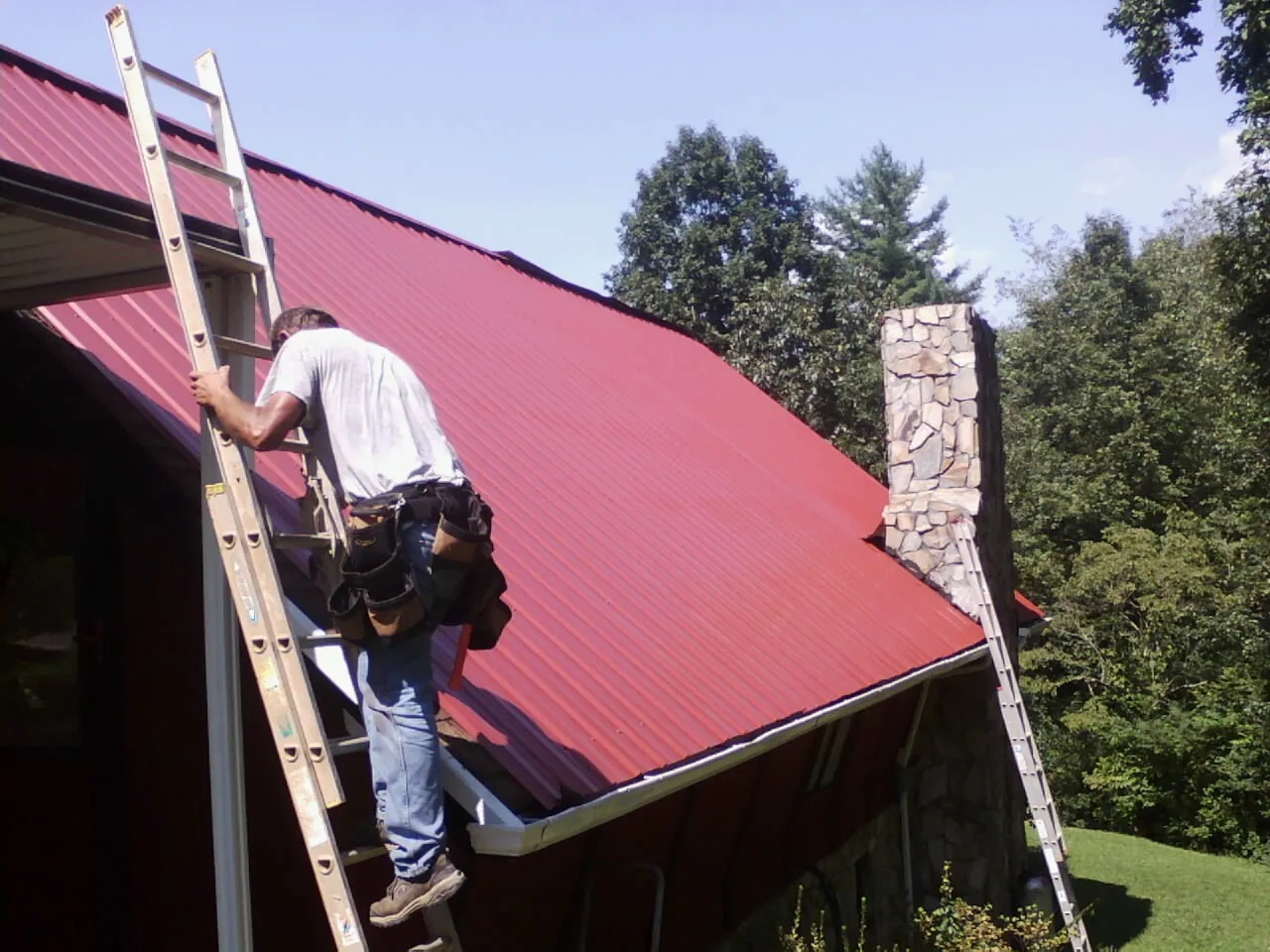 Roofer on a ladder beside a red metal roof and stone chimney — Soffit & Fascia Repair by Roofing Brothers in Loganton